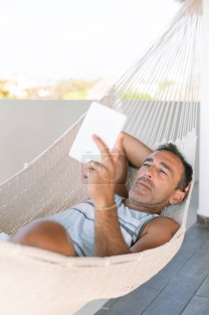 Photo for A man has nestled into a comfortable hammock on a luxurious rooftop terrace. While enjoying the fresh air at heights, he immerses himself in his e-book during vacation. High quality photo - Royalty Free Image