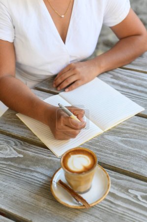 Téléchargez les photos : Angle élevé de culture femme anonyme portant une blouse blanche prenant des notes dans le planificateur tout en étant assis à la table en bois avec une tasse de latte aromatique pendant la pause café - en image libre de droit