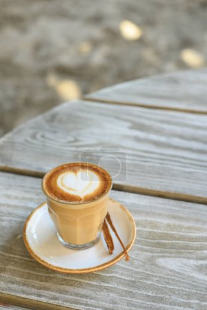 Foto de Alto ángulo de café aromático en taza de vidrio con latte arte en forma de corazón colocado en platillo con palo de vainilla - Imagen libre de derechos