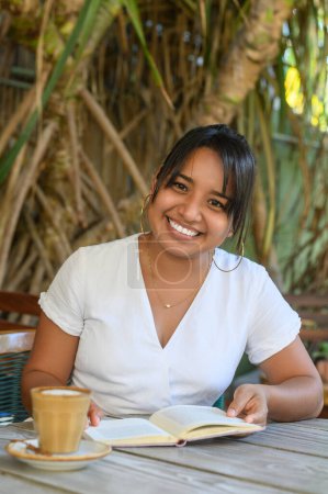 Foto de Joven hembra india positiva sentada a la mesa con taza de café con leche aromática mientras lee un interesante cuento en un libro descansando en una cafetería al aire libre - Imagen libre de derechos