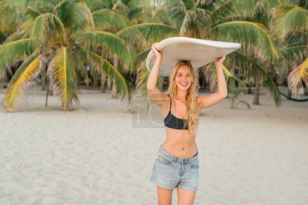 Photo for Delighted young female traveler in black top and denim shorts with blond hair smiling at camera while carrying surfboard on head against tropical palms - Royalty Free Image