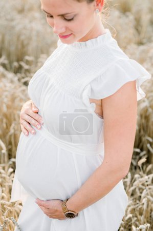 Photo for Crop young pregnant female in white dress touching tummy while standing in grassy field in daylight and looking down against blurred dried grass - Royalty Free Image