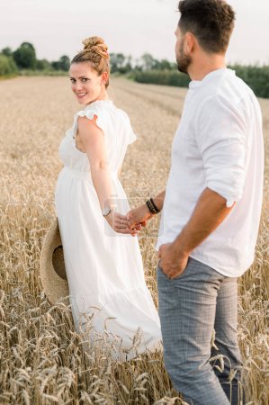 Photo for Side view of young pregnant woman in white white dress holding hand of husband while walking in grain field and looking at each other - Royalty Free Image