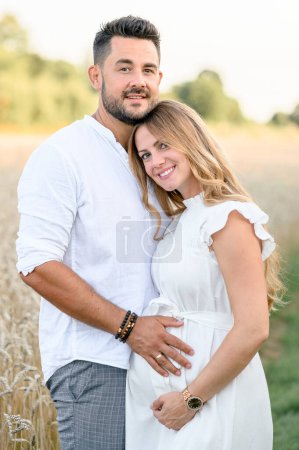 Foto de Mujer embarazada joven en vestido romántico blanco abrazándose con el hombre mientras está de pie con las manos en el vientre en el campo de hierba y mirando a la cámara - Imagen libre de derechos