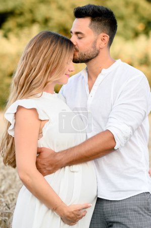 Photo for Side view of young pregnant woman in white dress hugging and kissing husband with closed eyes while standing in countryside on blurred filed background - Royalty Free Image