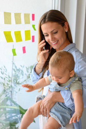 Téléchargez les photos : Une jeune mère positive et un bébé mignon en tenue décontractée souriant et ayant une conversation téléphonique tout en se tenant près de la fenêtre avec de nombreuses notes collantes colorées à la maison - en image libre de droit
