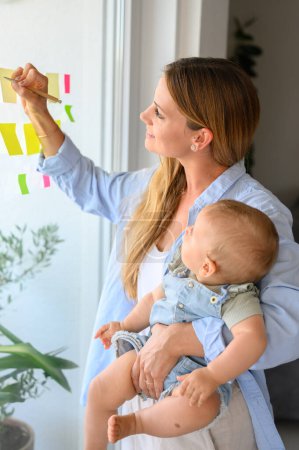 Foto de Concentrado joven madre en ropa casual de pie cerca de la ventana con el pequeño bebé y escribir información sobre notas adhesivas en casa - Imagen libre de derechos