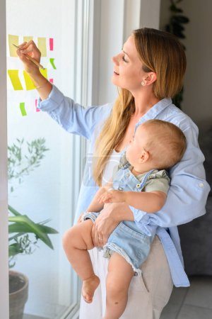 Photo for Positive young mother and adorable little baby in casual wear smiling and taking notes on sticky papers while standing near window at home - Royalty Free Image