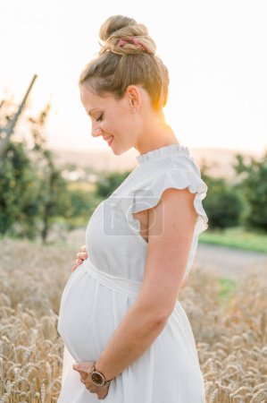 Photo for Side view of young pregnant female in white dress looking down and touching belly while standing in field in sunlight against blurred landscape - Royalty Free Image