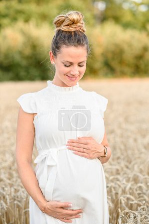Téléchargez les photos : Souriant jeune femme prénatale en robe blanche regardant vers le bas tout en touchant la bosse de bébé avec les mains et debout dans un champ herbeux par une journée ensoleillée à la campagne - en image libre de droit