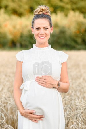 Photo for Positive young expecting female in white dress smiling and looking at camera while standing in field with dry grass and touching tummy with hands against blurred nature - Royalty Free Image
