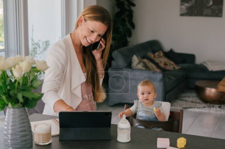 Photo for Delighted young mother with long hair smiling and talking on smartphone while using tablet near adorable baby girl during remote work from home - Royalty Free Image