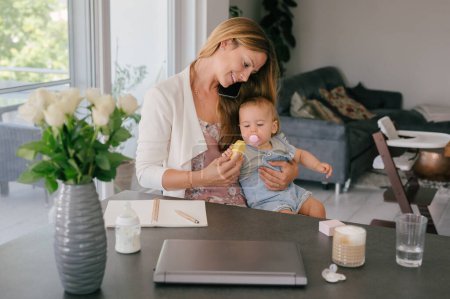 Photo for Loving young mother with long hair smiling and caressing adorable infant daughter while sitting at table with notebook and laptop during distance work at home - Royalty Free Image