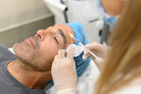 Foto de De arriba del paciente masculino que recibe la inyección facial que rejuvenece de la esteticista femenina de la cosecha en la clínica médica estética - Imagen libre de derechos