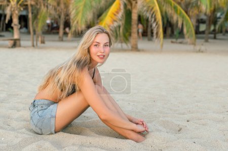 Photo for Full body side view of slim young blonde tourist in beach clothes sitting on sand and looking at camera while bending forward against palm trees - Royalty Free Image