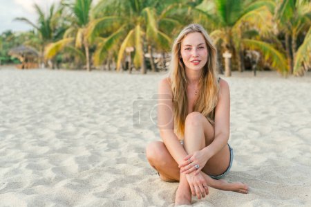 Foto de Cuerpo completo de alegre joven rubia viajera femenina sentada en la orilla del mar de arena y sonriendo mientras mira a la cámara contra las palmas exóticas durante las vacaciones de verano - Imagen libre de derechos
