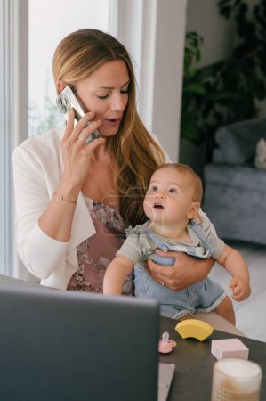 Foto de Mujer positiva con lindo niño pequeño en regazo sentado en la mesa con el ordenador portátil y hablando en el teléfono móvil mientras trabaja desde casa - Imagen libre de derechos