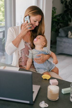 Foto de Joven madre independiente hablando en el teléfono inteligente mientras sostiene al bebé lindo y se sienta en la mesa con el ordenador portátil y los juguetes en la sala de estar en casa - Imagen libre de derechos
