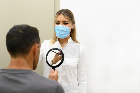 Téléchargez les photos : Focalisé jeune femme cosmétologue debout dans le marquage de masque avec crayon sur le visage d'un patient masculin méconnaissable avec réflexion dans le miroir tenu à la main tout en regardant vers le bas dans la clinique cosmétique moderne - en image libre de droit