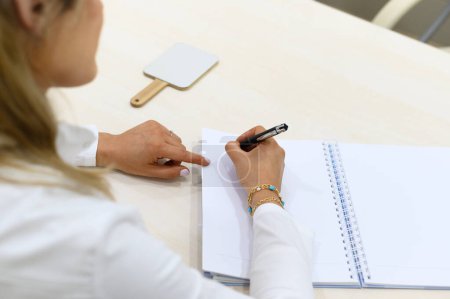 Photo for High angle of unrecognizable female consultant sitting at wooden table with handheld mirror and looking down while taking notes in notebook with pen in cosmetic clinic - Royalty Free Image