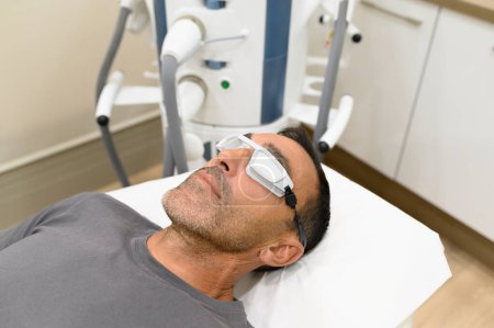 Foto de Alto ángulo de paciente varón adulto acostado en la cama con gafas de protección láser en la habitación con equipo mientras se prepara para el procedimiento de tratamiento en la clínica moderna - Imagen libre de derechos
