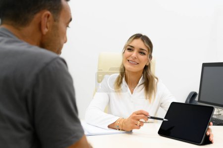 Téléchargez les photos : Cosmétologue souriante assise à table avec écran d'ordinateur et montrant la tablette à un patient masculin méconnaissable tout en se regardant et en expliquant les détails du traitement en clinique - en image libre de droit