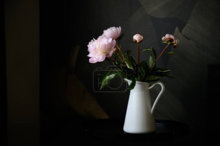 Photo for Composition of white ceramic jug with fresh blooming flowers placed on table in dark room - Royalty Free Image