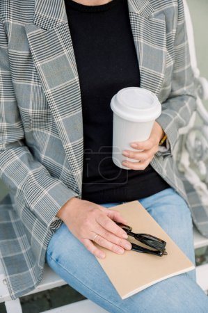 Téléchargez les photos : Cultivez une femme anonyme en tenue tendance assise à table avec une tasse de café en papier et tenant un carnet en papier et des lunettes de soleil le jour. - en image libre de droit