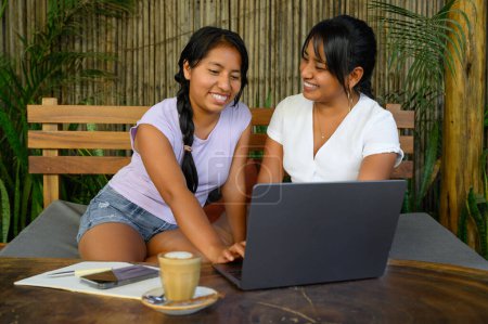 Téléchargez les photos : Positif jeune couple asiatique en vêtements décontractés assis à la table avec ordinateur portable et tasse de café tout en souriant et en se regardant - en image libre de droit