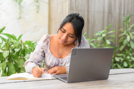 Téléchargez les photos : Jeune Mexicaine en tenue décontractée assise à table avec ordinateur portable et prenant des notes dans un carnet tout en travaillant sur un projet à distance - en image libre de droit