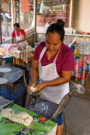 Foto de Cocinera mexicana amasando masa y rodando en bola cerca de prensa de tortilla con envoltura de plástico mientras hace sabroso pan plano chapati desde cero - Imagen libre de derechos