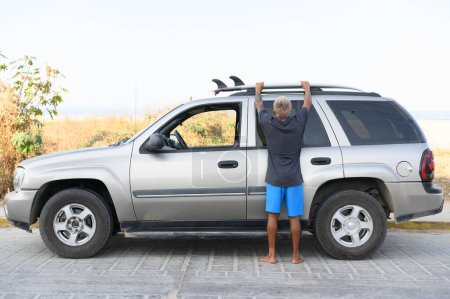 Photo for Back view of crop anonymous surfer in casual clothes standing on pavement near modern car and attaching surfboard to the roof rack on van. travel content - Royalty Free Image