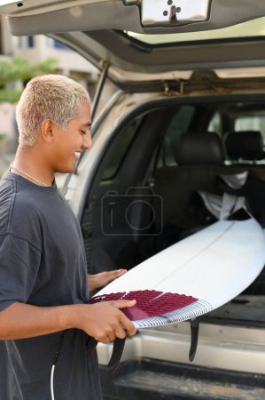 Téléchargez les photos : Vue latérale du jeune Mexicain aux cheveux blonds en vêtements décontractés se tenant près de la voiture et mettant la planche de surf en voiture après avoir surfé sur la plage - en image libre de droit