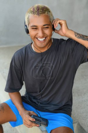 Téléchargez les photos : Jeune homme latino positif aux cheveux blonds en chemise noire et short souriant et regardant la caméra assis dans la rue - en image libre de droit