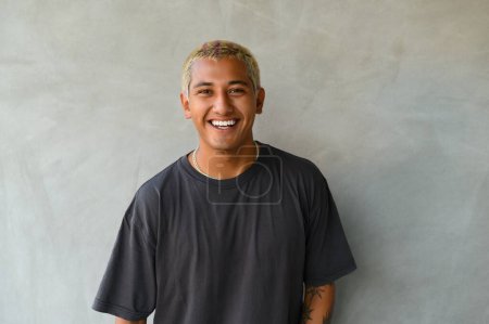 Foto de Joven hombre positivo en camisa negra sonriendo y mirando a la cámara mientras está de pie sobre fondo gris - Imagen libre de derechos