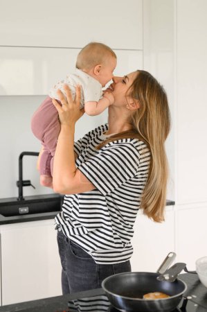 Photo for Side view of young mother and happy little baby girl in casual clothes standing in kitchen, Mother throws baby up, laughs and plays. family concept - Royalty Free Image