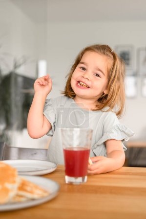 Téléchargez les photos : Petite fille mignonne en vêtements décontractés souriant et regardant la caméra tout en étant assis à table avec une tasse de boisson et une délicieuse crêpe dans la cuisine - en image libre de droit