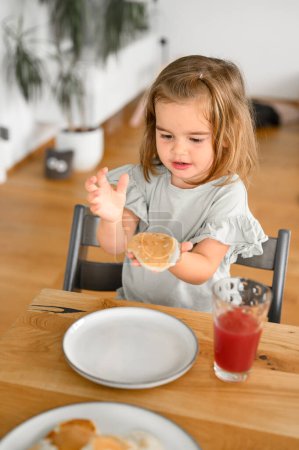 Foto de Linda niña sentada en la mesa de madera y comiendo delicioso panqueque de desayuno en la mañana en casa. concepto familiar - Imagen libre de derechos