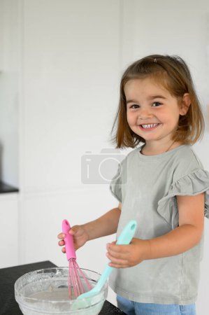 Téléchargez les photos : Petite fille mignonne en vêtements décontractés debout dans la cuisine, mélangeant la pâte pour des crêpes fraîches et regardant la caméra. concept de famille - en image libre de droit
