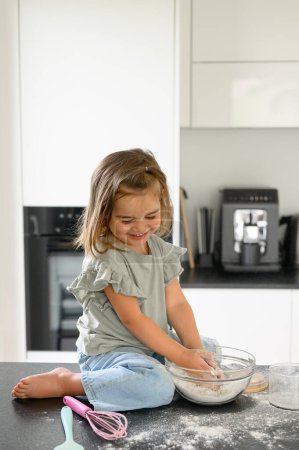 Photo for Little smiling girl in casual clothes sitting in kitchen counter and preparing cookies in kitchen at home. family concept - Royalty Free Image