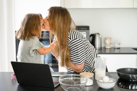 Téléchargez les photos : Vue latérale de la jeune mère caucasienne embrasser fille tout en faisant des biscuits frais faits maison dans la cuisine moderne à la maison s - en image libre de droit