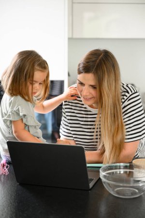 Téléchargez les photos : Jeune femme avec une petite fille en vêtements décontractés souriant et regardant l'écran d'ordinateur portable tout en recherchant des recettes dans la cuisine à la maison - en image libre de droit