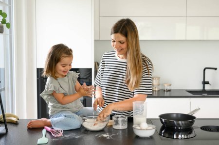 Photo for Side view of mother and daughter in casual clothes standing at modern kitchen counter and preparing dough for making pancakes in kitchen at home. family concept - Royalty Free Image