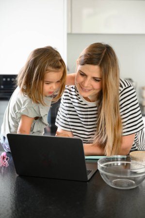 Foto de Las mujeres jóvenes con hija en ropa casual sentado a la mesa con el ordenador portátil, la búsqueda y la receta de cocina en el ordenador portátil en la cocina en casa - Imagen libre de derechos