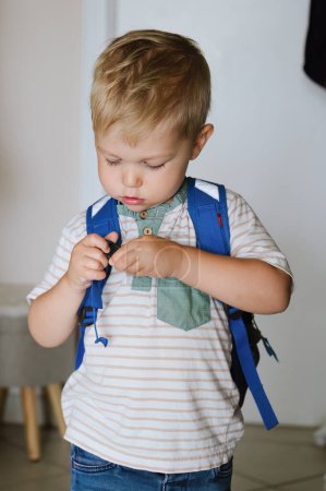 Foto de Adorable niño pequeño en ropa casual de pie en la habitación de luz y ponerse la mochila antes de ir a la escuela - Imagen libre de derechos