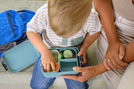 Photo for Cropped image of blond-haired boy and unrecognizable female holding container of food after kindergarten. back to school. homemade meals. High quality photo - Royalty Free Image