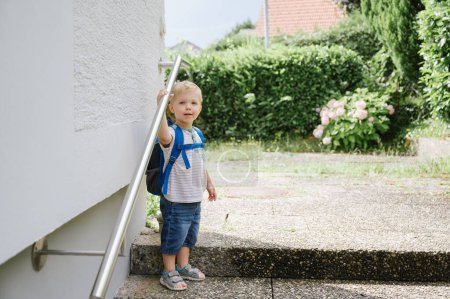 Téléchargez les photos : Vue latérale du petit garçon joyeux portant un jean bleu et une chemise rayée debout près de l'escalier et regardant la caméra par une journée ensoleillée à l'extérieur - en image libre de droit