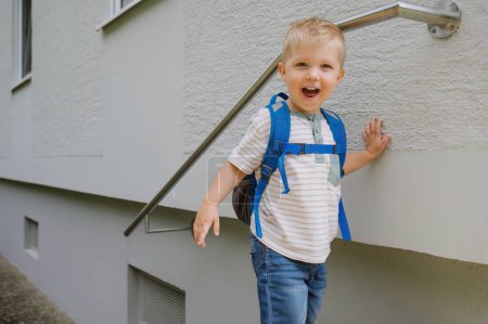 Téléchargez les photos : Vue de face de l'adorable tout-petit garçon debout près de l'escalier et souriant à la caméra tout en portant un sac à dos. - en image libre de droit
