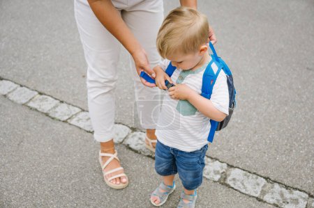 Téléchargez les photos : Maman en vêtements décontractés aider son petit fils aux cheveux blonds et tenue d'été pour mettre un sac à dos avant d'aller à l'école. concept de retour à l'école - en image libre de droit