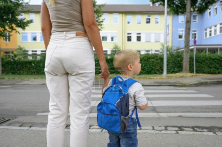 Photo for Anonymous woman in casual clothes holding backpack in hand and toddler son crossing the street on the crosswalk. little boy looking to the right side. back to school concept - Royalty Free Image